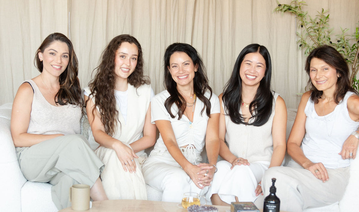 Five women sitting together on a white sofa in a bright, modern room with beige curtains, a light wood coffee table, and a patterned rug.